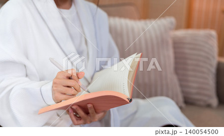 adult women writing morning journal in notebook wearing white bathrobe sitting on couch at home relaxing planning daily routine mindfulness practice self care 140054500