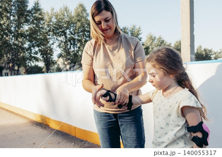 woman helps young girl put on roller skate protective gear. They are outdoors, enjoying quality time together. family bonding and activity 140055317