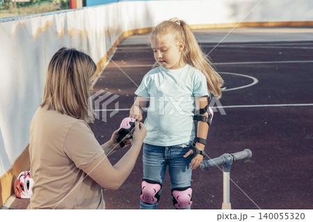 woman helps young girl put on roller skate protective gear. They are outdoors, enjoying quality time together. scene reflects family bonding and activity woman helps young girl put on roller skate protective gear. They are outdoors, enjoying quality time together. scene reflects family bonding and activity 140055320