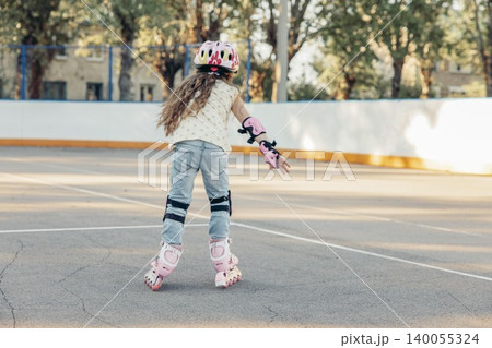 young Caucasian girl with long brown hair skates on a smooth surface. She wears a pink helmet and protective gear, enjoying quality time outdoors. Back view young Caucasian girl with long brown hair skates on a smooth surface. She wears a pink helmet and protective gear, enjoying quality time outdoors. Back view 140055324
