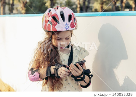 girl with long brown hair wears a pink helmet and protective gear. She stands by wall using a smartphone girl with long brown hair wears a pink helmet and protective gear. She stands by wall using a smartphone 140055329