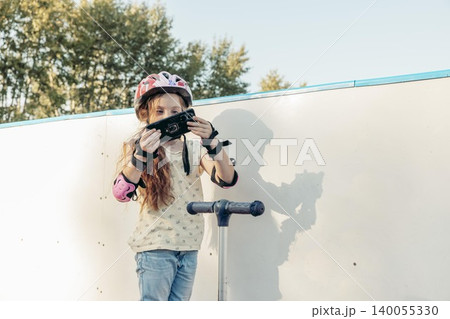 girl with long brown hair wears pink helmet and protective gear. She stands by wall using a smartphone 140055330