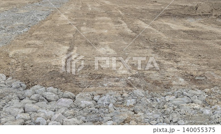A construction scene with large gray rocks gravel in the foreground. The background is compacted brown earth soil displaying clear tire tracks from heavy machinery. A construction scene with large gray rocks gravel in the foreground. The background is compacted brown earth soil displaying clear tire tracks from heavy machinery. 140055375