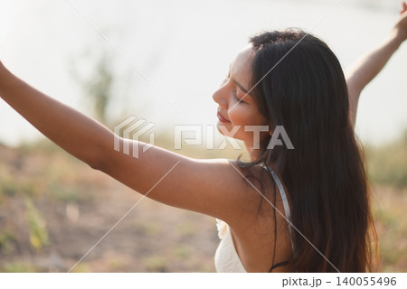 Peaceful woman in white lace dress stretching arms in soft lakeside sunlight, eyes closed and smiling, expressing fresh air, summer freedom, mindfulness, nature wellness, and relaxed outdoor joy. 140055496