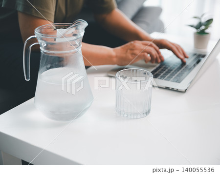 Glass water pitcher and tumbler on white desk with blurred person typing on laptop, showing hydration, wellness, remote work, healthy office routine, productivity, and modern home workspace. Glass water pitcher and tumbler on white desk with blurred person typing on laptop, showing hydration, wellness, remote work, healthy office routine, productivity, and modern home workspace. 140055536
