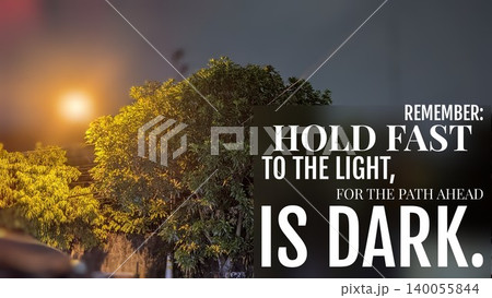 A tree's silhouette is cast against a bright street light at night, featuring the quote, "HOLD FAST TO THE LIGHT, FOR THE PATH AHEAD IS DARK." This image inspires perseverance hope through difficulty. 140055844