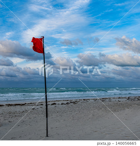 Red warning flag standing tall on a beach with cloudy skies and waves 140056665