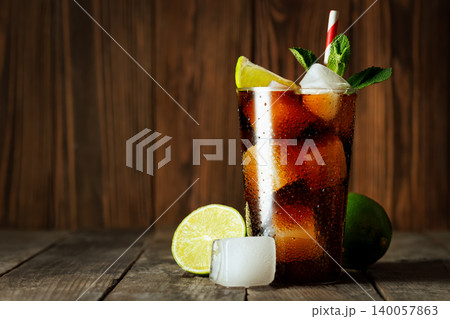 Cuba libre sparkling cola and rum cocktail is chilling in glass with condensation, ice, lime slices and fresh mint on weathered wooden surface Cuba libre sparkling cola and rum cocktail is chilling in glass with condensation, ice, lime slices and fresh mint on weathered wooden surface 140057863
