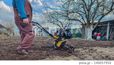 Man operating a walk-behind tractor, tilling and cultivating spring garden soil in a backyard plot on a cloudy day, preparing ground for planting and homestead work. Man operating a walk-behind tractor, tilling and cultivating spring garden soil in a backyard plot on a cloudy day, preparing ground for planting and homestead work. 140057958