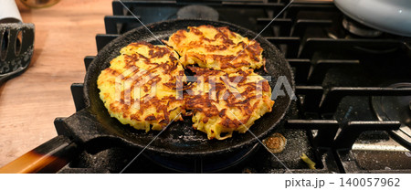 Crispy golden brown potato pancakes frying in a hot cast iron pan on a gas stove burner, preparing a delicious homemade meal or snack in a kitchen setting. Crispy golden brown potato pancakes frying in a hot cast iron pan on a gas stove burner, preparing a delicious homemade meal or snack in a kitchen setting. 140057962