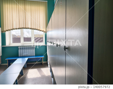 Empty changing room featuring a row of light wooden lockers, blue benches, and a window with blinds, creating a clean and functional space for students or athletes. Empty changing room featuring a row of light wooden lockers, blue benches, and a window with blinds, creating a clean and functional space for students or athletes. 140057972