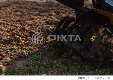 Walk-behind tractor tilling garden soil with rugged blades, preparing earth for planting and new crops; close-up of rototiller working rich brown soil in spring sunlight.. Walk-behind tractor tilling garden soil with rugged blades, preparing earth for planting and new crops; close-up of rototiller working rich brown soil in spring sunlight.. 140057980