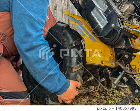 Farmer replacing a large, knobby tire on a yellow walk-behind tractor, focusing on the maintenance and repair of gardening and farming equipment for soil cultivation. Farmer replacing a large, knobby tire on a yellow walk-behind tractor, focusing on the maintenance and repair of gardening and farming equipment for soil cultivation. 140057995