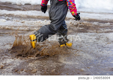 Child wearing colorful waterproof clothing and yellow rubber boots jumping and splashing in a dirty, muddy puddle outdoors on a wet, snowy day, creating dynamic motion and joyful play. Child wearing colorful waterproof clothing and yellow rubber boots jumping and splashing in a dirty, muddy puddle outdoors on a wet, snowy day, creating dynamic motion and joyful play. 140058004