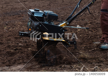 Man operating a walk-behind tractor, cultivating and preparing the soil for planting in a home garden, demonstrating spring gardening work and agricultural practices. Man operating a walk-behind tractor, cultivating and preparing the soil for planting in a home garden, demonstrating spring gardening work and agricultural practices. 140058009