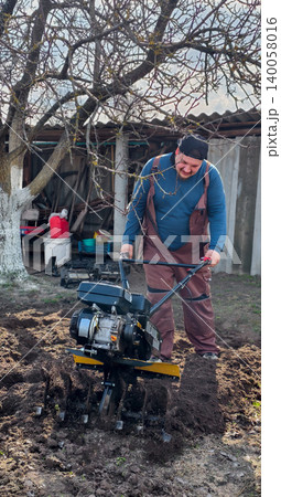 Man plowing garden soil with a walk-behind tractor, cultivating the ground in spring for planting, working outdoors in the backyard to prepare for agricultural endeavors. Man plowing garden soil with a walk-behind tractor, cultivating the ground in spring for planting, working outdoors in the backyard to prepare for agricultural endeavors. 140058016