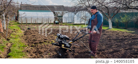 Man working in the garden, operating a walk-behind tractor to cultivate the soil. Greenhouses are visible in the background, showing a scene of active farm work and preparing the ground for planting. 140058105