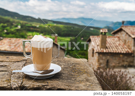 Foamy latte coffee on rustic stone terrace with countryside village backdrop Foamy latte coffee on rustic stone terrace with countryside village backdrop 140058197