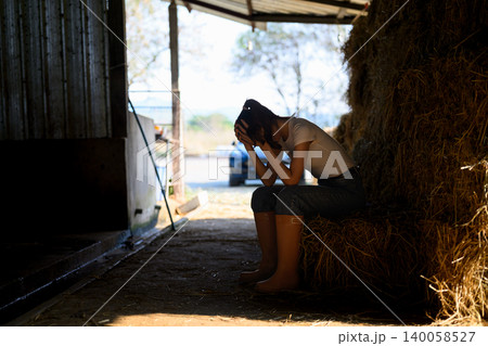 Exhausted female farm worker sitting on hay bale in barn feeling stressed and overwhelmed Exhausted female farm worker sitting on hay bale in barn feeling stressed and overwhelmed 140058527