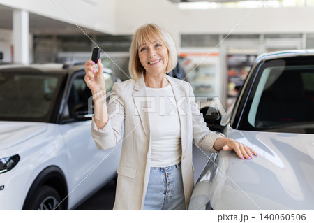 Smiling senior woman holding car key, standing inside modern dealership showroom 140060506