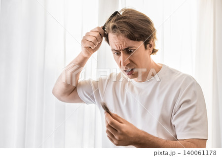 Shocked male grooming with a comb looks at hair loss in his hand. Concept of a man dealing with stress, alopecia, and early baldness. Shocked male grooming with a comb looks at hair loss in his hand. Concept of a man dealing with stress, alopecia, and early baldness. 140061178