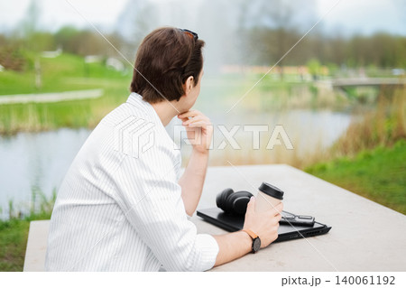 Back view of a freelance man relaxing in an outdoor park. He is taking a break from laptop work, holding a coffee and thinking while looking at nature. 140061192