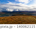 A wide landscape view from a grassy hill overlooking a valley with rolling hills, green fields, and forests under a partly cloudy blue sky. 140062115