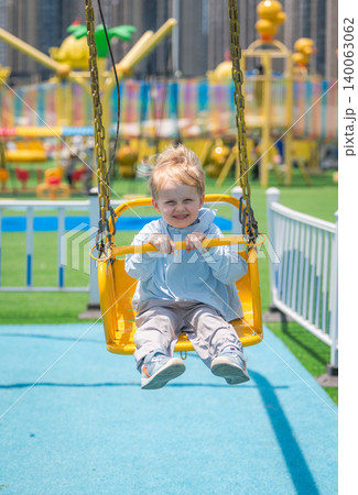 A cheerful blond boy smiles while swinging high on a yellow swing at a brightly colored amusement park. 140063062