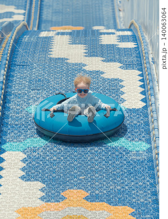 Blonde child in sunglasses rides a blue inflatable tube down an outdoor dry slide on a bright day Blonde child in sunglasses rides a blue inflatable tube down an outdoor dry slide on a bright day 140063064