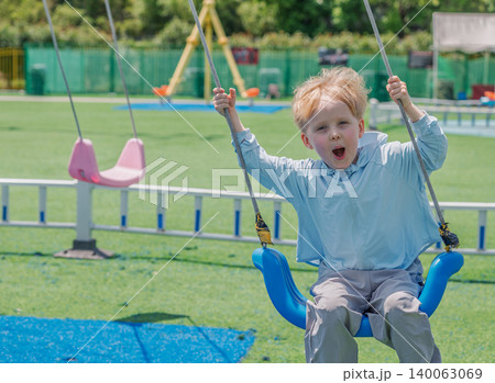 Excited young blonde boy swings high on a blue swing at a bright green outdoor playground on a sunny day Excited young blonde boy swings high on a blue swing at a bright green outdoor playground on a sunny day 140063069