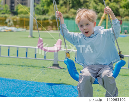 Excited young blonde boy swings high on a blue swing at a bright green outdoor playground on a sunny day Excited young blonde boy swings high on a blue swing at a bright green outdoor playground on a sunny day 140063102