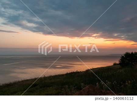 Vast water surface reflects vibrant sunset sky from a grassy hillside with trees during evening Vast water surface reflects vibrant sunset sky from a grassy hillside with trees during evening 140063112