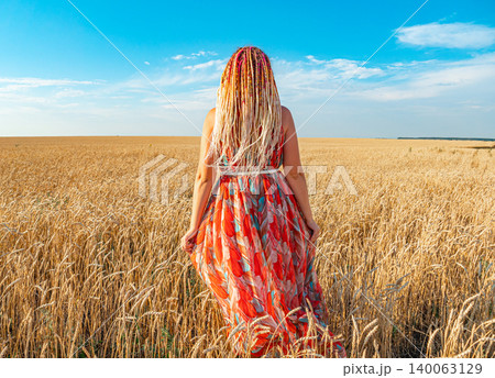 Woman in patterned dress with long braided hair stands in golden wheat field on sunny summer day 140063129