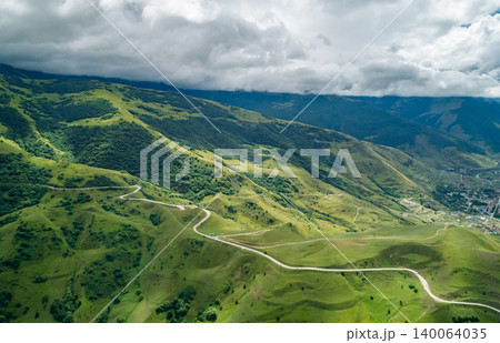 Panoramic view of verdant hills and valleys crossed by a winding road under a dramatic cloudy sky in a mountain range Panoramic view of verdant hills and valleys crossed by a winding road under a dramatic cloudy sky in a mountain range 140064035