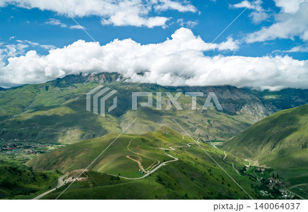 Panoramic view of serpentine roads wind through lush green mountains where clouds meet peaks and a valley village rests below 140064037