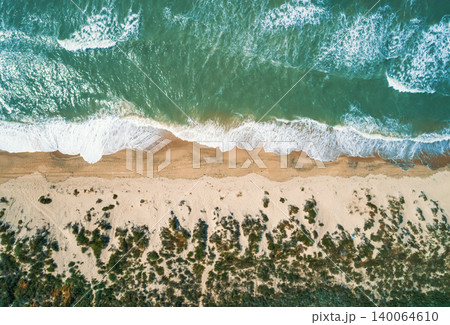Aerial view shows vibrant green ocean water with white foam waves crashing onto a tan sandy beach. Aerial view shows vibrant green ocean water with white foam waves crashing onto a tan sandy beach. 140064610