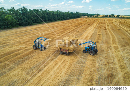 Farmers working together to collect hay bales using tractors in a sunlit field during late summer 140064630