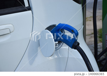 Close-up view of an electric vehicle charging port with a blue connector inserted into a white car, showcasing the charging mechanism and the vehicle's bodywork in detail 140066365