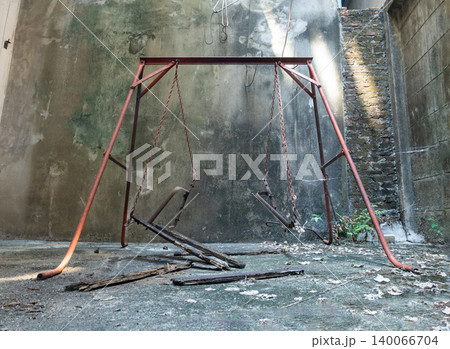 A weathered, crimson swing set stands as a solitary monument to forgotten laughter against a stained, monolithic concrete wall. Time and neglect have dismantled its purpose, leaving the chains A weathered, crimson swing set stands as a solitary monument to forgotten laughter against a stained, monolithic concrete wall. Time and neglect have dismantled its purpose, leaving the chains 140066704