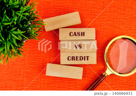 Close up view of wooden blocks spelling Cash vs Credit on a vibrant orange background with a green plant and magnifying glass, representing financial decisions Close up view of wooden blocks spelling Cash vs Credit on a vibrant orange background with a green plant and magnifying glass, representing financial decisions 140066931