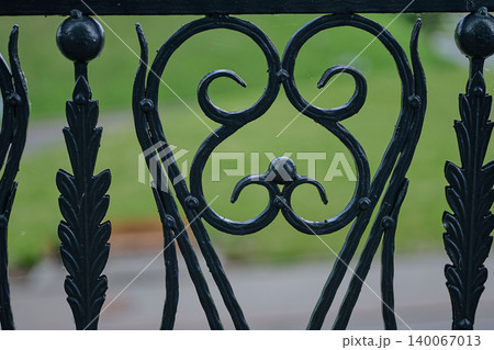 Close-up captures ornamental dark ironwork of a fence panel. Heart-shaped scrolls blend with leafy designs, set against a soft green outdoor landscape during daylight hours 140067013