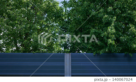 Dense green leaves of two large trees extend above a dark blue corrugated sound barrier with a grey post. Bright blue sky appears through the foliage on a clear day 140067024