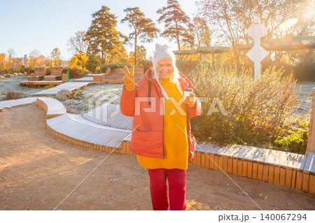 woman with coffee cup showing victory sign in city park on frosty autumn morning. Caucasian lady enjoy cold sunny day outdoors. Lifestyle portrait of mature female in warm clothes. woman with coffee cup showing victory sign in city park on frosty autumn morning. Caucasian lady enjoy cold sunny day outdoors. Lifestyle portrait of mature female in warm clothes. 140067294