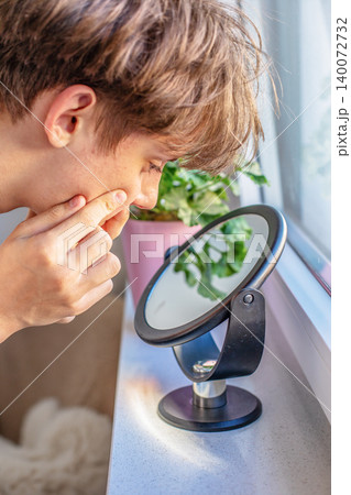 Young caucasian teenager guy looks into mirror and squeezes acne pimple on his face. Skin problem, dermatology care and youth health concept Young caucasian teenager guy looks into mirror and squeezes acne pimple on his face. Skin problem, dermatology care and youth health concept 140072732