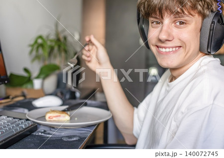 Smiling teenage gamer wearing headset and eating sweet pastry from a plate while sitting at the PC desk at home. Smiling teenage gamer wearing headset and eating sweet pastry from a plate while sitting at the PC desk at home. 140072745