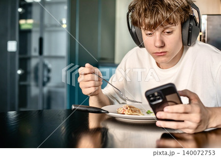 Focused teenage boy wearing headphones uses his smartphone while having a meal at the kitchen table, distracted by digital content. Focused teenage boy wearing headphones uses his smartphone while having a meal at the kitchen table, distracted by digital content. 140072753