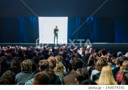 Large audience watching speaker deliver presentation on illuminated stage at conference with focused crowd energy 140074591