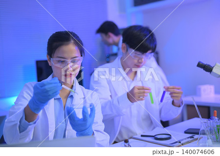 Female scientist using a pipette while colleagues compare green and purple samples, showing teamwork and precision in modern laboratory research Female scientist using a pipette while colleagues compare green and purple samples, showing teamwork and precision in modern laboratory research 140074606