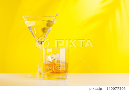 Glass of whiskey with ice and martini with olives on yellow background 140077303