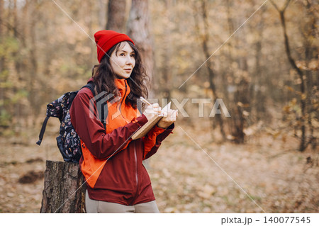 Woman stands in autumn forest while writing in notebook and looking away. Wilderness survival curriculum, nature-based learning program, forest school activities. 140077545